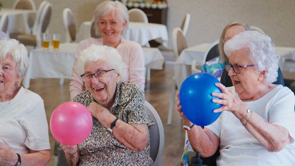 Three senior residents and a caregiver smiling together in a bright, comfortable communal living room.