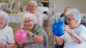 Three senior residents and a caregiver smiling together in a bright, comfortable communal living room.